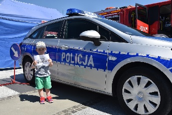 Mały chłopiec stojący na tle radiowozu policyjnego trzyma w ręku kolorowankę