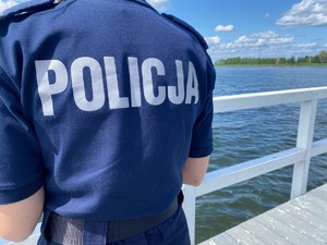 Policjant odwrócony tyłem stoi na pomoście akwenu wodnego.