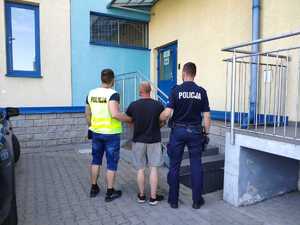 dwaj  policjanci prowadzą zatrzymanego. Jeden z policjantów ma na sobie kamizelkę z napisem Policja, drugi jest w policyjnym mundurze