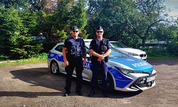 dwaj policjanci stoją przy radiowozie, w tle drzewa