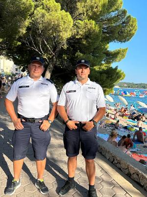 dwaj polscy policjanci stoją na promenadzie nadmorskiej, w tle na plaży wypoczywający ludzie