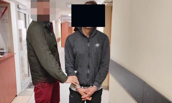 Nieumundurowany policjant stoi obok i trzyma za nadgarstek mężczyznę, który ma założone kajdanki na ręce trzymane z przodu