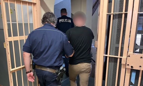 dwaj umundurowani policjanci prowadzą zatrzymanego, z boku widoczne są okratowane drzwi, które są otwarte