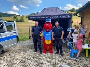 Dwaj policjanci pozują do zdjęcia z maskotką policyjną po ich prawej stronie uczestnicy pikniku.