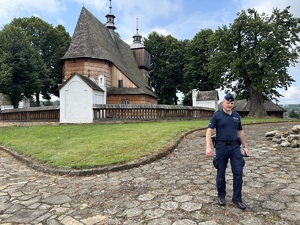 na pierwszym planie widać umundurowanego policjanta, który stoi przed kościołem w miejscowości Blizne, będący jednocześnie zabytkiem wpisanym na światową listę UNESCO. W tle zdjęcia widoczna zabudowa sakralna z otaczającym kościół murem oraz sam budynek kościelny