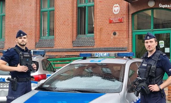 Zdjęcie przedstawia dwóch umundurowanych policjantów na tle radiowozu służbowego