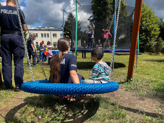 widok z tyłu: policjantka z dzieckiem siedzą na huśtawce, przy nich z boku widać stojącego policjanta. Przed nimi widać trampolinę, na której skaczą dzieci