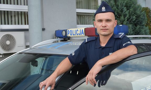 Zdjęcie kolorowe. Umundurowany policjant na tle radiowozu