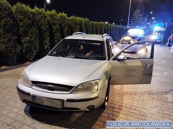 Pojazd osobowy marki Ford koloru srebrnego za nim stoi policyjny radiowóz zdjęcie wykonane nocą