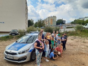 Policjant pozuje do zdjęcia z grupą dzieci, obok stoi radiowóz.
