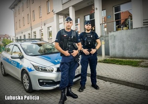 dwaj umundurowani policjanci stojący przy radiowozie, w tle budynek komendy Policji