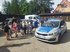 Policjant stoi obok radiowozu pokazując grupie dzieci jego wnętrze.