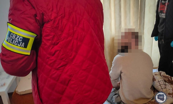 Wnętrze pokoju. Zatrzymany mężczyzna siedzi na łóżku. Obok stoją funkcjonariusze CBZC