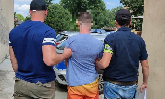 Dwaj policjanci w cywilnych ubraniach prowadzą zatrzymanego mężczyznę skutego w kajdanki do radiowozu