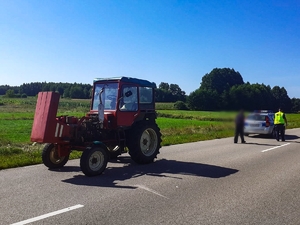 Na drodze stoi traktor z podniesioną maską, za nim stoi radiowóz policyjny, w którego stronę idzie policjant i mężczyzna.