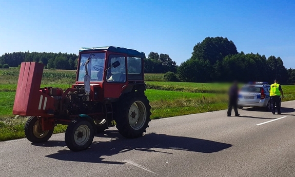 Na drodze stoi traktor z podniesioną maską, za nim stoi radiowóz policyjny, w którego stronę idzie policjant i mężczyzna.