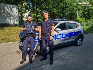 Zdjęcie dwóch policjantów, którzy stoją obok oznakowanego radiowozu