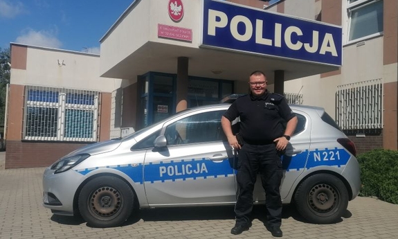 policjant stoi przy radiowozie policyjnym w tle budynek komisariatu Policji