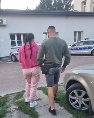 Zdjęcie kolorowe, policjant prowadzi zatrzymaną kobietę ubraną na różowo. Z przodu przed budynkiem zaparkowane dwa radiowozy.