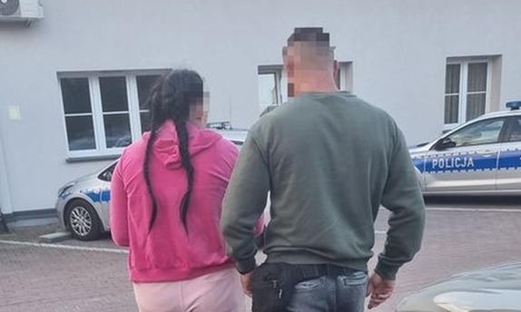 Zdjęcie kolorowe, policjant prowadzi zatrzymaną kobietę ubraną na różowo. Z przodu przed budynkiem zaparkowane dwa radiowozy.