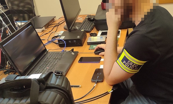 policjant siedzi przy biurku przy laptopie, na biurku leżą dwa smartfony i nośniki pamięci