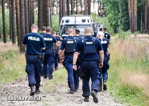 grupa umundurowanych policjantów idzie leśną drogą, przed nimi stoi policyjny radiowóz