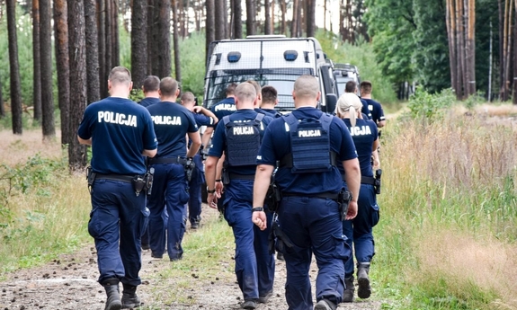 grupa umundurowanych policjantów idzie leśną drogą, przed nimi stoi policyjny radiowóz