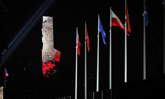 Pora nocna. Podświetlony na biało-czerwono pomnik na Westerplatte. Po prawej stronie flagi Polski, Unii europejskiej i Miasta Gdańska na masztach.