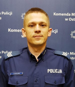 umundurowany policjant stoi na tle baneru Komendy Miejskiej Policji w Ostrołęce