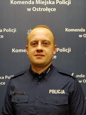 umundurowany policjant stoi na tle baneru Komendy Miejskiej Policji w Ostrołęce