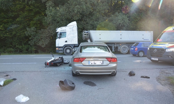 Na zdjęciu pojazd marki audi, z jego lewej stroni na jezdni roztrzaskany motocykl, z pawiej karetka pogotowia. W oddali jadący drugim pasem ruchu samochód ciężarowy. W tle las