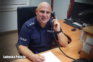 Policjant oficer dyżurny siedzi przy swoim stanowisku służby, trzyma słuchawkę telefonu przy uchu