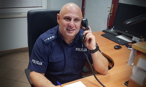 Policjant oficer dyżurny siedzi przy swoim stanowisku służby, trzyma słuchawkę telefonu przy uchu