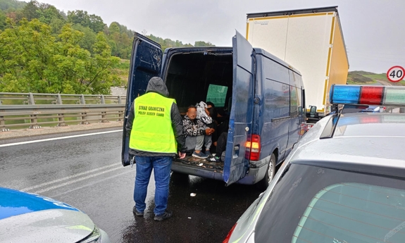 fragment radiowozów w tle otwarty bus, w środku którego siedzą osoby obok stoi funkcjonariusz straży granicznej