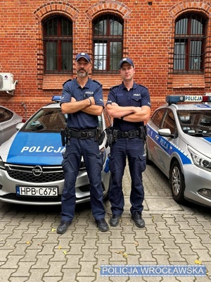 Dwaj umundurowani policjanci stojący na tle zaparkowanych radiowozów i budynku z cegły