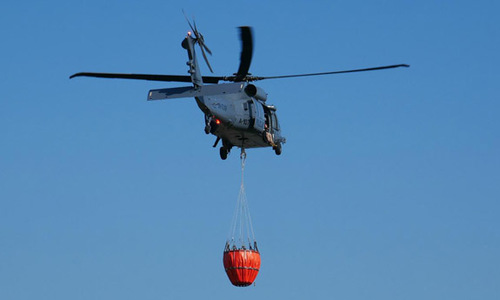 Policyjny śmigłowiec w locie z podwieszonym zbiornikiem na wodę.