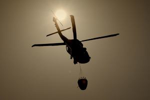Policyjny śmigłowiec w trakcie lotu z podwieszonym zbiornikiem na wodę na tle zachodzącego słońca.