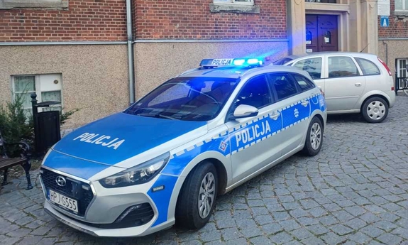radiowóz z włączonymi sygnałami świetlnymi stoi przed szpitalem, za nim stoi inny samochód