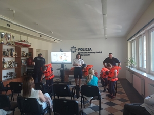 dzieci ubrane w kamizelki ratunkowe w kolorze pomarańczowym, policjanci stojący pomiędzy dziećmi szczegółowo instruują jak należy właściwie użytkować sprzęt ratunkowy. Opiekunowie siedzą na krzesłach oglądając prezentację multimedialną.