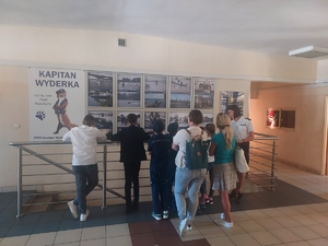 Laureaci gali odwiedzają Komisariat Rzeczny w Warszawie. Osoby stoją przy plakatach i zdjęciach tematycznie związanych z miejscem i słuchają policjantki, która opowiada o zadaniach policji wodnej.