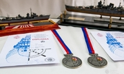 na pierwszym planie dwa medale i dyplomy leżące na stole, w tle dwa modele okrętów wojennych
