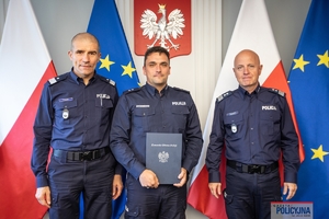 Komendant Główny Policji gen. insp. Jarosław Szymczyk i Zastępca Komendanta Głównego Policji nadinsp. Roman Kuster stoją z policjantem na tle flag Polski i Unii Europejskiej, na ścianie wisi godło Polski.