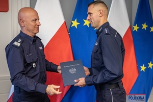 Komendant Główny Policji gen. insp. Jarosław Szymczyk z policjantem na tle flag Polski i Unii Europejskiej, na ścianie wisi godło Polski