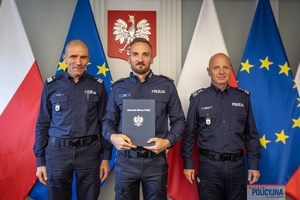 Komendant Główny Policji gen. insp. Jarosław Szymczyk i Zastępca Komendanta Głównego Policji nadinsp. Roman Kuster stoją z policjantem na tle flag Polski i Unii Europejskiej, na ścianie wisi godło Polski.