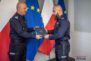 Komendant Główny Policji gen. insp. Jarosław Szymczyk z policjantem na tle flag Polski i Unii Europejskiej, na ścianie wisi godło Polski.