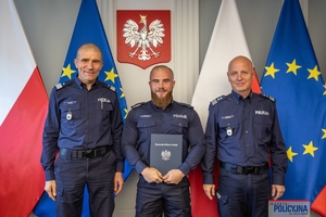 Komendant Główny Policji gen. insp. Jarosław Szymczyk i Zastępca Komendanta Głównego Policji nadinsp. Roman Kuster stoją z policjantem na tle flag Polski i Unii Europejskiej, na ścianie wisi godło Polski.