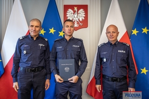 Komendant Główny Policji gen. insp. Jarosław Szymczyk i Zastępca Komendanta Głównego Policji nadinsp. Roman Kuster stoją z policjantem na tle flag Polski i Unii Europejskiej, na ścianie wisi godło Polski.