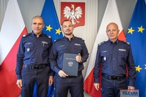 Komendant Główny Policji gen. insp. Jarosław Szymczyk i Zastępca Komendanta Głównego Policji nadinsp. Roman Kuster stoją z policjantem na tle flag Polski i Unii Europejskiej, na ścianie wisi godło Polski.