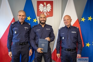 Komendant Główny Policji gen. insp. Jarosław Szymczyk i Zastępca Komendanta Głównego Policji nadinsp. Roman Kuster stoją z policjantem na tle flag Polski i Unii Europejskiej, na ścianie wisi godło Polski.