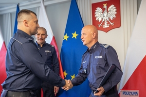 Komendant Główny Policji gen. insp. Jarosław Szymczyk i Zastępca Komendanta Głównego Policji nadinsp. Roman Kuster stoją z policjantem na tle flag Polski i Unii Europejskiej, na ścianie wisi godło Polski.